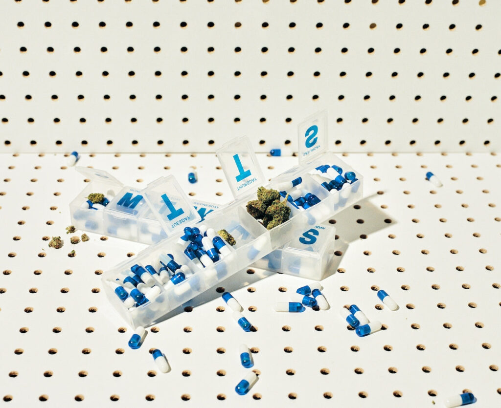 Close-up of a weekly pill box containing blue and white capsules, with some compartments holding cannabis buds, set against a perforated background.