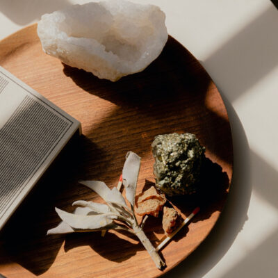 A flat lay image of a crystal and a sage bundle laying on a round wooden tray with shadows overcasting | Unearthing the Historical and Cultural Origins of Modern-Day Rituals | ritual origin, examples of modern-day rituals | Sugar Magnolia