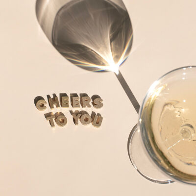 Flat lay image of a champagne coupe glass casting a shadow, with the words “Cheers to You” displayed in sparkly gold letters on a white background.