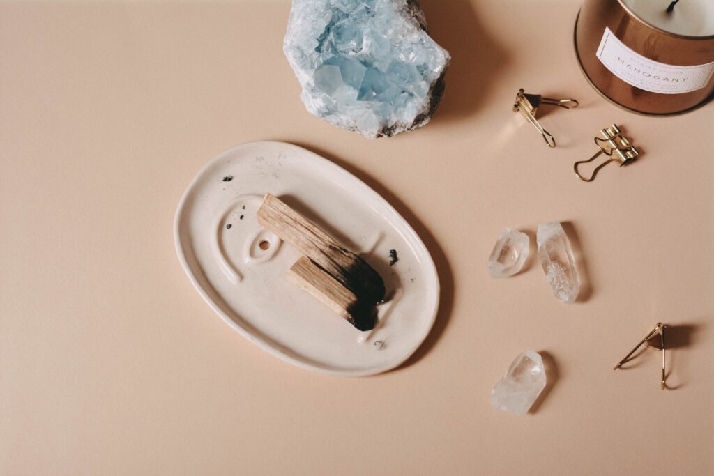 A smudge stick of Palo Santo placed on a white ceramic dish, surrounded by quartz crystals, a large blue crystal, and a lit candle.