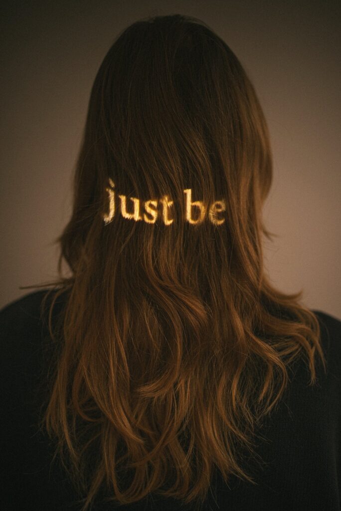 The back of a woman’s head with long brown hair, illuminated by the projected words “just be” in golden light, symbolising mindfulness and presence.