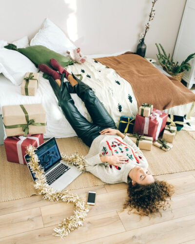 A woman laying on her bedroom floor with her feet on her bed and one hand on her chest with Christmas presents and a laptop surrounding her