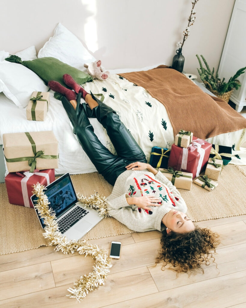Woman in a festive sweater lying on the bedroom floor beside a bed, surrounded by wrapped Christmas gifts, a laptop, and a phone.