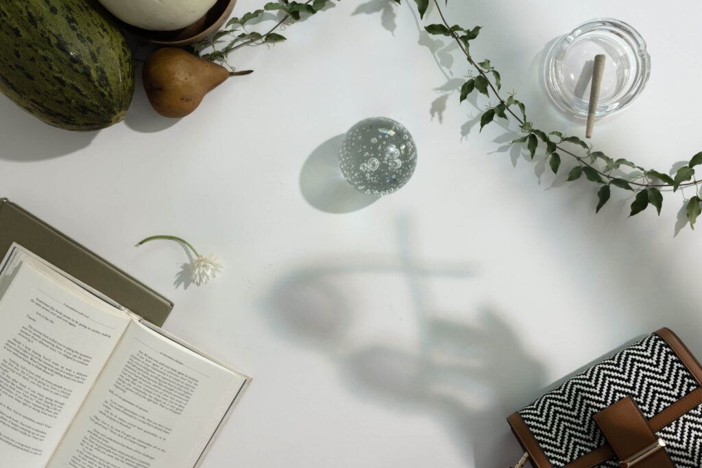 Flat lay image of self-care ritual essentials including a joint in an ashtray, a clear crystal ball, a daisy flower, an open book, and a patterned pouch surrounded by green leaves on a white background
