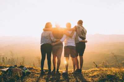 Group of friends standing arm in arm on a hilltop, watching the sunset over the mountains.