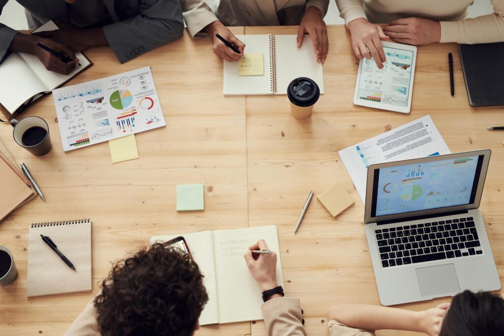 Group of people working around a wooden table with laptops, reports, charts, and notebooks.