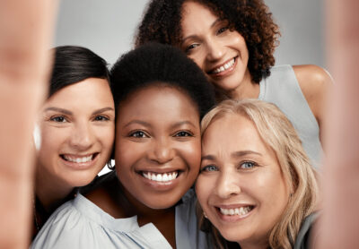 Group of four happy, diverse women smiling while taking a selfie together, celebrating connection and emotional wellness.