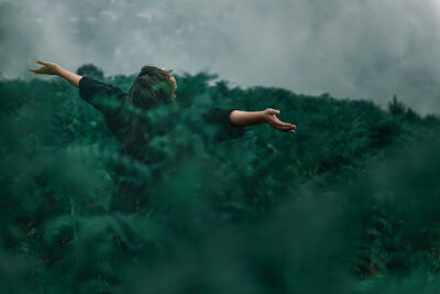 Woman walking freely through lush green forest, arms outstretched, surrounded by nature’s calm.