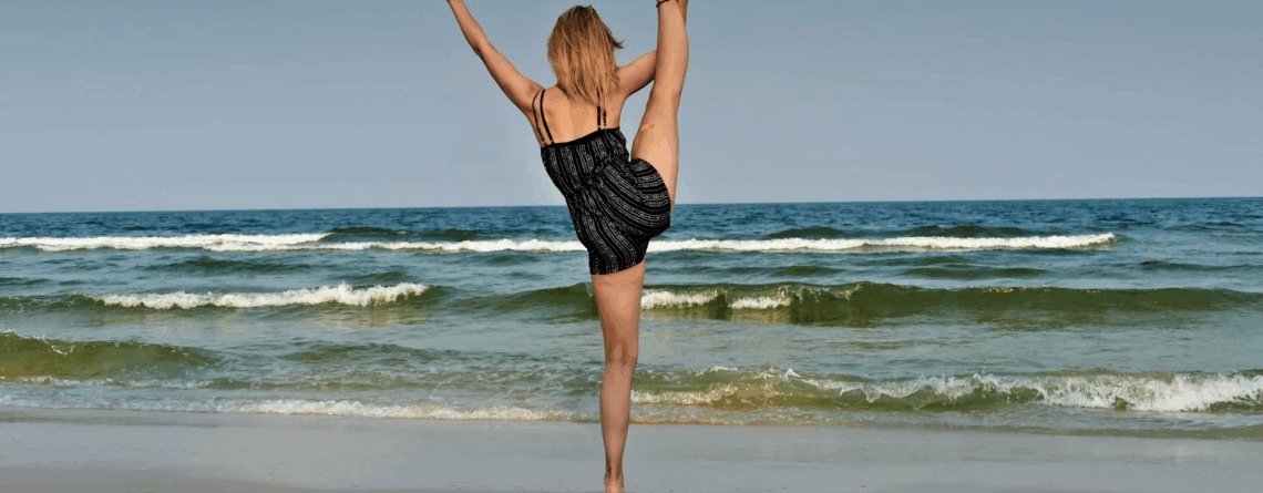 Woman stretching with high leg lift on the beach, facing the ocean